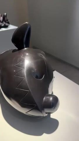 The video shows a shiny, black, abstract sculpture that resembles a stylized animal figure, possibly a rabbit or dog. The sculpture has simplified geometric shapes and a smooth, reflective surface. It is sitting on a white surface. The camera slowly pans around the sculpture, showing its shape and details from different angles, including the bottom. The room appears to be a gallery or display space, with neutral-colored walls.