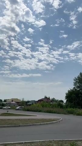 The video shows a suburban landscape with a blue sky filled with scattered white and gray clouds. A paved road curves through the foreground. In the background, several houses with red roofs are visible, surrounded by green trees and vegetation. Two cars are parked on the left side of the road. The sun is partially obscured by clouds, creating variations in light and shadow across the sky. The camera is positioned low, looking upwards at the sky.