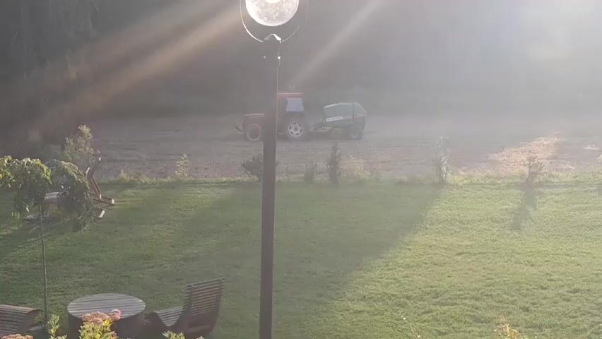 This media file captures a rural scene in Bukowina Tatrzańska, Poland, during what appears to be early morning or late afternoon due to the strong, angled sunlight casting long shadows and creating a hazy atmosphere. The primary subjects are agricultural. A red tractor with a green baler attachment is actively working in a field, suggesting haymaking or a similar harvesting operation. The field itself is a muted brown and tan, with visible rows indicating recent activity.

In the foreground, a well-maintained grassy lawn dominates the lower half of the frame. A round wooden table with chairs is visible, along with a piece of outdoor furniture, suggesting a residential area adjacent to the field. A tall, dark pole with a light fixture stands centrally in the foreground, a man-made object within the natural landscape. Scattered trees and shrubs dot the scene, with denser foliage visible in the background, hinting at a wooded area beyond the cultivated field.

The weather appears to be clear but hazy, possibly due to dust from the tractor's work or atmospheric conditions typical of dawn or dusk. The strong sunlight and its illuminating effect on the haze create a somewhat ethereal or tranquil mood. There are no people visible in the frame, emphasizing the focus on the agricultural machinery and the landscape. No text is discernible in the media file. The overall impression is one of rural productivity and the quiet beauty of a working agricultural landscape bathed in soft light.