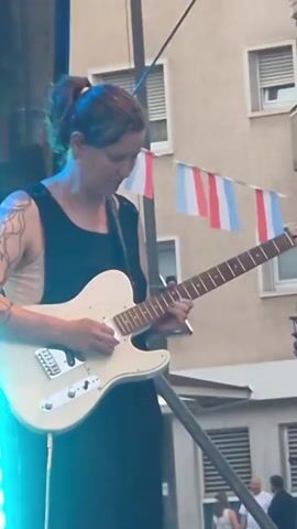 A person is playing a white electric guitar on a stage. They are wearing a black sleeveless dress and have a tattoo on their arm. There are blue stage lights to the left of the person. In the background, there is a building with windows, and some flags are hanging. Another person is visible on the left side of the stage playing a bass guitar. There are also some sunflowers decorating the stage.