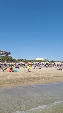 A wide shot shows a sandy beach with numerous beachgoers relaxing on lounge chairs under umbrellas. The water is calm and clear. In the foreground, the edge of the water laps onto the sand. A person is standing on the sand in the left of the frame, tossing a ball upwards. Further down the beach, several people are in the water, some swimming and others wading. A breakwater made of large rocks extends into the sea. The sky is a clear, bright blue.
