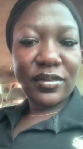 This is a close-up video of a woman in what appears to be the interior of a vehicle, possibly a car or bus, given the presence of headrests in the background. The woman is wearing a black top and a black headscarf. She has on makeup, including mascara and dark lipstick. A thin gold necklace is visible around her neck.

The video captures the woman from the chest up, with her face as the primary focus. She displays a range of emotions throughout the short clip, starting with a neutral or slightly displeased expression, then transitioning to a smile, and finally a broader, more joyous smile. Her eyes are dark and expressive. There are some subtle skin imperfections visible, suggesting a natural complexion.

The lighting in the video appears to be natural, coming from the side and slightly from the front, indicating it is daytime. The background is slightly blurred, but the headrests of the seats are discernible. There is no visible text in the video. The overall impression is a personal and candid recording, likely a selfie video. The location context provided is Laminga, Nigeria.