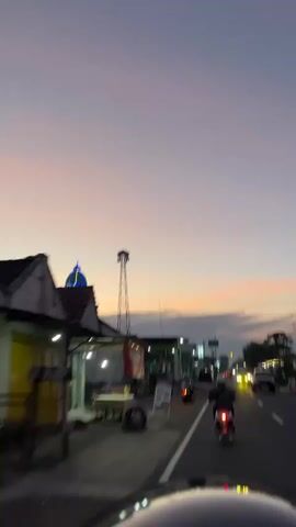 This media file captures a serene sunset scene in Purworejo, Indonesia. The sky transitions through soft hues of pink and purple, indicating dusk. A prominent mosque, adorned with green and blue accents, stands to the left, its dome illuminated. Beside it, a tall, metal tower with antennas rises against the colorful sky.

The scene depicts a street with a few motorbikes and a car, suggesting a moderately busy, yet peaceful, evening. Two individuals are seen riding motorbikes, their forms silhouetted against the fading light. The overall atmosphere is tranquil and somewhat contemplative, amplified by the soft twilight.

On the left side of the road, a building with a red and white striped awning is visible, possibly a shop or food stall. A sign with text is placed in front of it. Further down the street, another building with green and white architecture is visible. Streetlights are on, casting a warm glow on the road and the surrounding structures.

The presence of the mosque, the tower, and the general architecture suggest a location within a town or city in Indonesia. The activity appears to be the daily commute or people going about their evening routines as the day concludes. The weather seems calm, with no signs of strong winds or rain. The mood evoked is one of quiet beauty and the gentle transition from day to night in a local Indonesian setting.