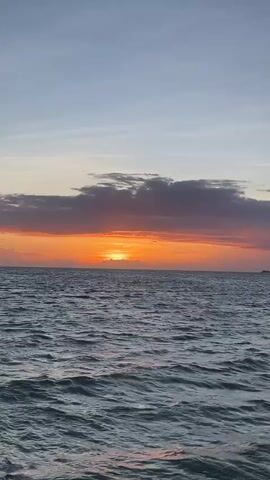 The video shows a sunset over the ocean. The sun is low on the horizon, casting a warm orange glow across the sky. There are some clouds in the sky, which are tinged with pink and purple. The ocean is choppy, with small waves. In the distance, there are two kitesurfers in the water, silhouetted against the sunset. There are also some smaller figures in the water, possibly other kitesurfers or swimmers. The video is shot from a low angle, giving the impression of being on a boat or the water.