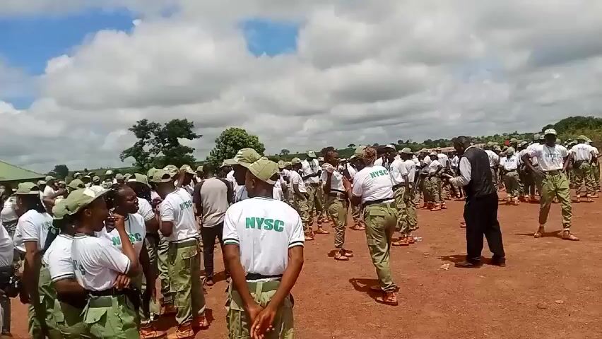 A medium shot shows a group of people wearing white shirts with "NYSC" printed on them and green camouflage pants, standing on a reddish-brown dirt field. The individuals are mostly facing different directions and appear to be in a casual gathering. Some are talking to each other, while others are looking around. In the background, more people wearing similar outfits can be seen, suggesting a larger group. A man in a black suit is also visible, seemingly interacting with some of the people. The background features a few scattered trees, a building with a green roof, and a cloudy sky. The camera moves slightly, panning across the scene, capturing more of the crowd and the surrounding environment.