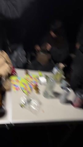 The video shows a group of young people gathered around a white folding table outdoors at night. On the table are several opened cans and bottles of alcoholic beverages, a container of lime juice, snack items, and confetti. A woman with long dark hair is sitting in a chair, holding a small dog in her lap. Another woman with red hair is standing by the table, playfully wrestling with someone out of frame. One person is seen with a green sour straw in their mouth. The area is dimly lit, making it difficult to discern specific details.