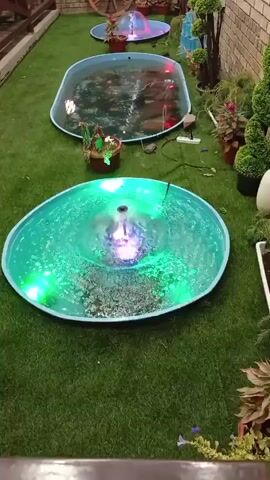 The video displays an outdoor setting with artificial turf flooring. Several circular shallow ponds are arranged in a line, each featuring a water fountain illuminated by colored LED lights. The closest pond has blue and pink lights, the middle pond exhibits green lights, and the furthest pond shows red and blue lights. Decorative elements like plants and a wooden wagon wheel are visible in the background. The ponds appear to be decorative features, possibly in a garden or patio area.