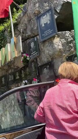 The video shows a memorial wall built into a rocky hillside, featuring numerous black plaques with photographs and names of individuals. A woman in a pink striped shirt is seen placing a bouquet of flowers on one of the plaques.

The memorial is adorned with flags, including a prominent green flag with an open hand and stars, which is the flag of Abkhazia. Red flags are also visible higher up. Large golden letters spelling out a word, possibly "АБХАЗИЯ" or a related place name, are affixed to the rock face.

Each plaque displays a black and white portrait of a man, along with their name and birth and death dates, predominantly in Cyrillic script. Many of the death dates consistently show March 16, 1993, indicating a collective loss or significant event on that day. Examples of visible names include ГУЧЕВ МУХАДИНОВИЧ, ПАЦУЛИЯ МИХАИЛ ВЛАДИМИРОВИЧ, КЪЫПШИНА РУСЛАНБЕЙ АРТЮМ-ИПА, КАСЛАНЗИА ЭДУАРД КАРБЕ-ИПА, ШЬАБАЛКЪАЗ-ИПА РУСЛАН, and ИНАПШБА БОРИС ТЫЩБА-ИПА.

The setting appears to be outdoors during daytime, with ample sunlight illuminating the memorial. Green foliage and trees are visible above and around the rocks, suggesting a natural, possibly mountainous, environment. Part of a dark-colored car is seen in the foreground, with its window reflecting the memorial. The scene evokes a sense of solemn remembrance and tribute.