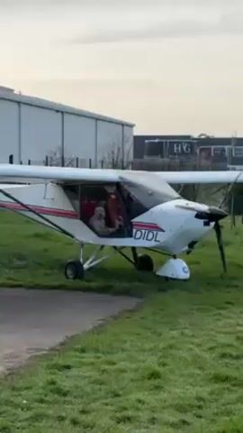 A small, white propeller airplane with the registration "DIDL" is parked on a patch of green grass next to a paved path.  The propeller is spinning, and there appears to be a dog sitting in the cockpit.  Behind the plane are large, industrial-looking buildings under a pale sky.  Some bare trees are visible.