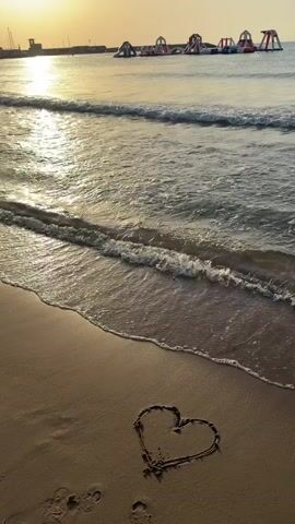 The video opens with a wide shot of a beach at sunrise or sunset. The sun is low on the horizon, casting a warm glow across the scene. In the foreground, waves gently lap against the sandy shore. A heart shape is drawn in the sand, facing the water. In the background, a series of inflatable water slides and structures are visible in the sea, suggesting a water park or recreational area. The water is calm, with gentle ripples and small waves. The overall impression is one of peace and tranquility.
