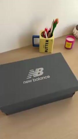 The video shows a person opening a New Balance shoe box on a light wooden surface. The box is grey with the New Balance logo on top. The person lifts the lid to reveal tissue paper with the New Balance logo printed all over. They then lift the tissue paper to reveal a pair of brown New Balance shoes. There are also several pairs of shoelaces in different colors: black and white, brown, and orange. A yellow mug labeled "BUTTER" with writing utensils inside is in the background, as well as a pink candle.