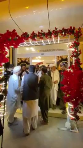The video captures a group of approximately 10-15 men gathered at a decorated entrance, likely an indoor venue, during what appears to be an evening event. The entrance is framed by an archway adorned with vibrant red artificial roses and illuminated by white string lights.

Most of the men are dressed in traditional attire, including light-colored shalwar kameez, some with dark waistcoats and turbans or other head coverings. They are engaged in greetings, shaking hands and exchanging embraces, suggesting a reception or arrival of guests. One prominent figure in a white shalwar kameez and white turban is seen interacting with others on the right side of the frame.

On the left, camera tripods and individuals appearing to be media personnel are visible, indicating that the event is being recorded or is of public interest. Through the partially open glass doors of the entrance, a brightly lit interior of the venue can be seen. The overall lighting within the scene is warm, casting a yellowish glow. The atmosphere appears to be social and cordial, consistent with a gathering in Kandahar, Afghanistan. No discernible text is visible.