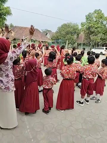 A group of young children in matching red and white uniforms are gathered outdoors, possibly at a school. Most of them are wearing red skirts or shorts and white shirts with red floral patterns. They appear to be participating in an activity, with many of them raising their hands in the air. An adult woman in a hijab and a patterned shirt is at the front, also with her hands raised. The background shows buildings, trees, and a clear sky. A white car is parked in the distance. The ground is paved with gray cobblestones.