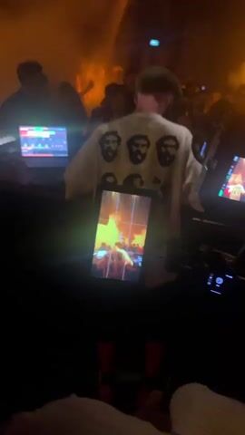 A person wearing a white t-shirt with black graphics is standing behind a DJ booth in a dimly lit club. The t-shirt has multiple circular graphics of faces on the back. The person is facing away from the camera, towards a crowd of people dancing. In front of the DJ booth, there are two laptops and DJ equipment. A tablet is mounted on a stand in front of the DJ, displaying an image. There is smoke or fog in the air, illuminated by stage lights, creating a vibrant atmosphere. The camera is positioned behind the person, capturing the scene from a low angle. The video is short, lasting only a few seconds.