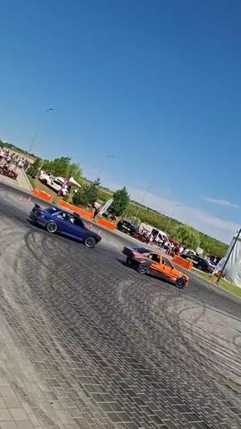 The video shows a sunny outdoor event with several cars performing drifts. The ground is paved with interlocking stones, and there are orange traffic cones placed around the course. In the background, there is a large white tent with some banners, and beyond that, a building and some trees. There are also spectators visible in the background, as well as some inflatable structures. The cars are the primary focus, with a blue car and an orange car performing drifts in the foreground. Another car, purple in color, is also visible and appears to be drifting as well. The scene suggests a car show or a drifting competition.