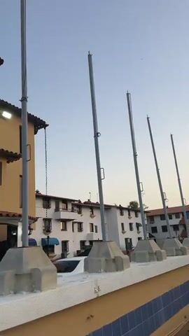 The video shows a street scene with various buildings and palm trees. The buildings are mostly two-story structures with a mix of white and yellow facades. Some buildings have balconies and traditional Spanish-style tiled roofs. Several metal flagpoles are visible, mounted on concrete bases along a low wall. The sky is a clear, pale blue, suggesting either early morning or late afternoon. There's a white car partially visible behind the wall. Palm trees are scattered throughout the scene.