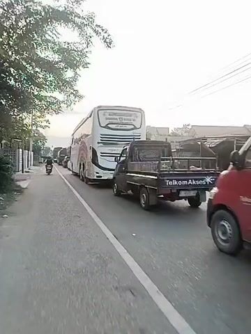 The video shows a large, white passenger bus with colorful rainbow-like stripes along its side, specifically red, orange, yellow, green, and blue. The bus is moving slowly in traffic on a paved road. The text "PUTRA PELANGI PERKASA" is visible on the side of the bus. A black pickup truck is seen in the foreground to the right of the bus, with the word "Telk" partially visible on its rear. Other vehicles, including cars and a motorcycle, are in the background, also in traffic. Trees and power lines line the side of the road. The sky is bright, suggesting daytime conditions. The camera appears to be moving alongside the bus. The setting is a road in Langa, Indonesia.