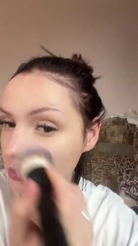 A close-up shot shows a young woman with her dark hair pulled up in a messy bun, framing her face. She has light skin with freckles and long eyelashes. She is wearing a white t-shirt. The woman is holding a makeup brush to her face, appearing to be applying powder or blush to her cheek and nose area. Her expression is neutral as she focuses on her makeup application. The background is blurred, but suggests an indoor setting with a textured wall and what looks like a patterned sofa. There are no visible text elements in the media. The lighting suggests daytime, but there are no specific indicators of the time of day or weather.