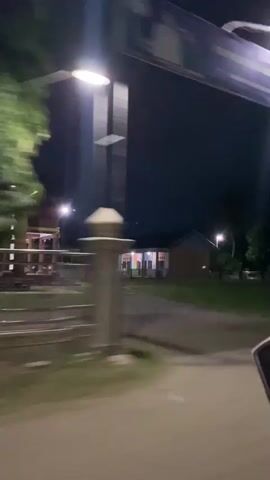 The video is a nighttime recording from a moving vehicle. The scene is dark, with streetlights illuminating some areas. There are buildings visible on the side of the road, with some lights on inside. Trees and vegetation are also present. The video is shaky due to the movement of the vehicle.