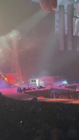 The video shows a live performance stage set bathed in red light and haze, featuring a central platform with an industrial backdrop including an oil rig structure and trailer. A figure in a white outfit stands prominently on the platform facing a large audience visible in the foreground. The stage is designed with rock-like formations. Above, a large spherical object hangs from the ceiling. The camera is handheld, capturing the scene from the audience's perspective. Throughout the clip, the lighting shifts between yellow and red hues.