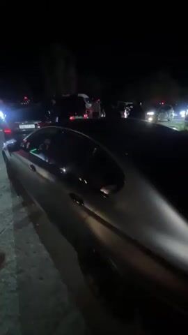 At night, the video shows a crowd of people and cars gathered on a paved road. In the foreground, a grey car with green interior lighting is parked. A woman in a white top and dark pants walks towards the back of the vehicle in front of it, which is black and has the license plate 06 TT 206. She briefly embraces a man wearing a black shirt and carrying a black bag, before the video ends. The area is dimly lit by car headlights and taillights. In the distance, more cars and people can be seen.