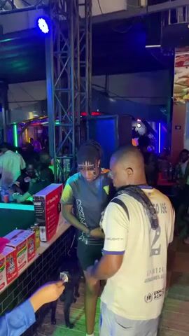 The video shows a man wearing a Barcelona soccer jersey standing near a tiled bar, where a box of "Indomie" instant noodles is visible. Another person approaches him, puts their arm around his shoulder, and whispers something to him. In the background, other people are seated at tables, and a few lights illuminate the scene. A dog is near the bar.