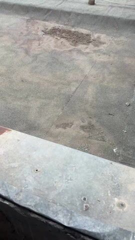 The video shows the roof of a building. The roof is flat and covered in a dark, tar-like material. There is a patch of damage in the center of the frame, where the tar is peeling away. The roof also has several small, square vents. A wire runs across the roof, anchored by small metal posts. In the background, there are trees and other buildings. The sky is cloudy.