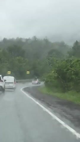 The video captures a view from inside a moving vehicle on a winding, wet road surrounded by dense green foliage. The road surface appears slick due to rain, with water visible on the asphalt. Several cars are traveling ahead, including a white SUV and a white van, both in the same lane, and a silver car further up the road. Yellow road signs indicating sharp turns are visible along the roadside. The sky is overcast and hazy, creating a muted atmosphere. The camera is positioned to give a sense of forward motion, with the scenery blurring slightly.