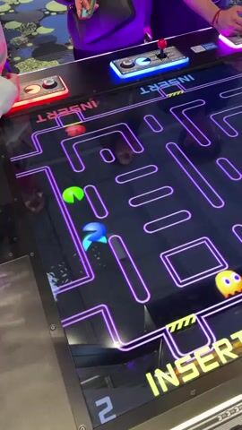Two individuals are actively engaged in a modern, oversized Pac-Man arcade game. The game features a large, horizontal screen displaying the iconic Pac-Man maze with neon purple lines. Pac-Man, along with several ghosts (yellow, light blue, and orange), are visible moving across the illuminated maze.

The game is designed for two players, with separate control panels positioned on opposite sides of the screen. Each panel includes an illuminated joystick and buttons, and one panel has the number "7450" visible. The players' hands are actively manipulating the controls, indicating they are in the midst of gameplay. One player wears a green wristband.

In the foreground, the words "INSERT COIN" are prominently displayed in bright yellow and green lettering on the game screen, suggesting the game is either waiting for payment or has just started/finished a round. The background shows glimpses of other arcade machines, contributing to the ambiance of an entertainment center or arcade. The overall lighting is dim, contrasting with the bright, vibrant colors emanating from the game's screen and controls, creating a lively and engaging atmosphere. The activity takes place in Schaumburg, United States of America.