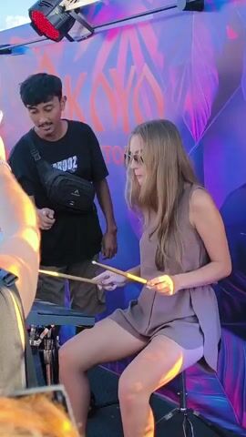 A woman is playing an electronic drum kit while sitting on a drum stool. She wears sunglasses and a sleeveless romper. A man stands behind her to her left. He's wearing a black t-shirt and khaki shorts with a small black bag across his torso. A colorful sign that reads “OKYO ROOFTOP” is directly behind them. Another person is partially visible in the lower left corner of the frame.