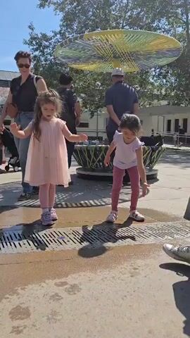 Two young girls play at the edge of a water feature on a sunny day. One wears a pale pink dress and matching tights, while the other wears a light purple shirt and pink leggings. They both have their arms extended, as if trying to catch the water. An adult woman with short dark hair, sunglasses, and jeans stands nearby, watching them. Several other adults and children are in the background, standing around a round structure with a colorful, umbrella-like decoration above it. The water feature is a rectangular grate with water trickling across it, creating puddles on the concrete surface. Trees and buildings can be seen in the background, suggesting a park or plaza setting.