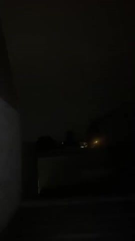 The video shows a dark outdoor scene at night. There is a brick wall on the left side. In the background, there are silhouetted buildings and trees. A faint yellow light is visible in the distance. A flash of light, likely lightning, illuminates the sky briefly around the 00:02 mark. The overall scene is dark and obscured by the nighttime conditions and possibly rain or fog.