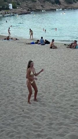 A young woman in a yellow bikini is on a sandy beach, holding a volleyball. She is looking up and preparing to hit the ball. In the background, other people are visible on the beach and in the water. The water is a clear blue, and there are some boats in the distance. The sky is overcast. The woman is in the center of the frame, and the focus is on her.