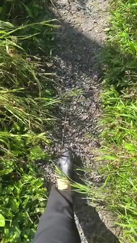 The video begins with a low-angle shot looking down at a gravel path. Green grass and foliage line both sides of the path. A person's dark-colored shoe and lower leg are visible in the foreground, indicating they are walking along the path. The camera perspective remains fixed, following the path as it continues forward. Small pieces of litter, such as a crumpled white wrapper, are visible on the path at various points throughout the clip. Shadows of branches and leaves from the surrounding vegetation intermittently fall across the path. The scene is brightly lit, suggesting it is daytime.