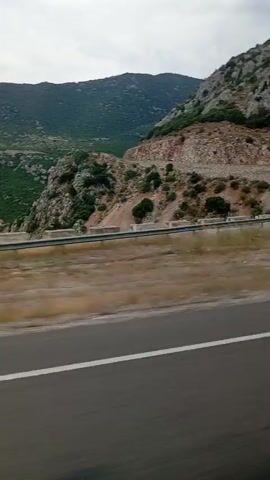 The video captures a scenic view of rolling hills and mountains from the perspective of a moving vehicle. The foreground shows a paved road with a white line, indicating it's a highway or a well-maintained road. Beyond the road, there's a dry, grassy embankment. The middle ground features more rugged terrain with patches of green vegetation scattered across rocky slopes. A metal guardrail runs along the edge of the road, separating it from the natural landscape. The background is dominated by large, verdant hills and mountains, with some areas appearing more sparsely vegetated and rocky. The sky is partly cloudy. The camera appears to be moving sideways or slightly backward, giving a dynamic sense of travel. There are no people or overt actions visible in the frame. The environment suggests a rural or mountainous region.