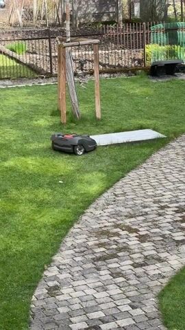 The scene unfolds on a bright, sunny day in a green backyard. A small, black robotic lawnmower with a red top sits on a patch of lush grass, next to a paved stone path. Behind the mower, there's a wooden structure consisting of two vertical posts and a slanted piece of wood. Further back, a dark metal fence separates the yard from a garden area with budding trees and shrubs. The person filming calls the lawnmower’s name and whistles, as if trying to get its attention.