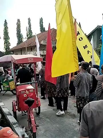 This video captures a lively street scene, likely a procession or celebration, in Sragen, Indonesia. A group of people, many dressed in traditional attire featuring striped tops and patterned bottoms, are moving forward. They are holding several colorful flags, predominantly bright yellow and red, with a few blue ones interspersed. One yellow flag prominently displays black lettering that reads "TOKO MAS," which translates to "Gold Shop" in Indonesian.

In the foreground, a red ice cream cart with the "KIMIA FARMA" logo is visible, suggesting a commercial aspect to the event or the presence of vendors. The people appear engaged and animated, hinting at a festive atmosphere. The time of day seems to be daytime, with diffused lighting suggesting it might be overcast or early morning/late afternoon. The setting appears to be a residential or commercial street lined with buildings and lush green trees, indicative of a warm climate. The presence of ice cream vendors and a gold shop suggests a local community event, possibly a parade, festival, or market day.