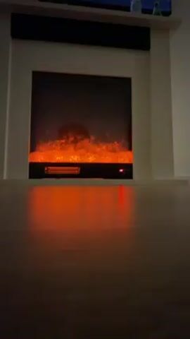 The camera is positioned at ground level and faces a white electric fireplace insert with a black frame. The fireplace is on, and the fake logs inside glow with orange light, creating the illusion of a burning fire. The light reflects onto the dark, shiny wooden floor in front of the fireplace. Above the fireplace is a white mantel with a black soundbar on top, and a television is visible above the mantel. Two bottles are also placed on the mantel, to the right. The room is dimly lit. The camera is stationary, and there is no movement or interaction in the frame. The focus is on the electric fireplace and its light.