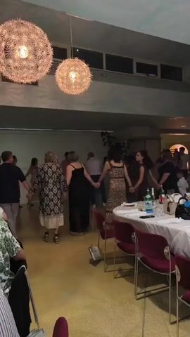 The video shows a group of people dancing in a circle in a room. There are two large spherical light fixtures hanging from the ceiling. Several tables are set up around the room with white tablecloths and chairs. The lighting in the room is dim with some spotlights illuminating the dancers. The people are dressed in casual to semi-formal attire. Some people are holding hands and moving in a coordinated dance. The floor appears to be made of a light-colored material. There is a wall with windows and some decorative elements on the right side of the frame. The overall atmosphere appears to be festive.