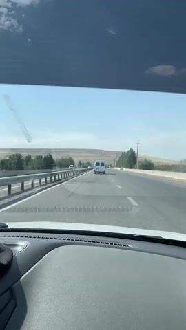 The video is shot from the perspective of a driver in a car, looking out through the windshield. The road ahead is asphalt, with visible cracks and patches. There are two lanes of traffic in the direction the car is moving. To the left of the road is a metal guardrail. Beyond the guardrail are trees and a dry, hilly landscape under a clear blue sky with scattered clouds. A white van is visible in the distance, driving in the same direction as the car. There are other vehicles on the road, but they are further away. The interior of the car, specifically the dashboard, is visible in the foreground. The lighting suggests it is daytime, likely sunny.