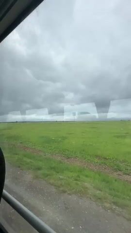 The video shows a view from inside a vehicle, looking out a window at a wide, flat, green field under a cloudy sky. The field appears to be agricultural land, with some rows of dirt visible. There are a few scattered trees or power poles in the distance. The foreground shows part of the vehicle's interior, including a seat and a window frame. The perspective suggests the vehicle is moving, as the landscape appears to be passing by.