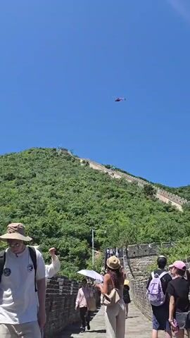 A red helicopter flies in a clear blue sky over a green, tree-covered mountain. Below the mountain, the Great Wall of China is visible, with a stone walkway where several people are standing, looking up at the helicopter. Some individuals are wearing hats and carrying bags, while others are holding umbrellas. The scene appears to be a tourist destination on a sunny day.
