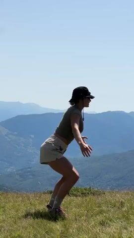 A woman wearing a hat and athletic clothing performs squats on a grassy hillside with a mountain range in the background. The video captures the woman in various stages of the squatting motion, from a deep squat with her arms extended forward to an upright position with her arms outstretched. The lighting suggests it is daytime with clear skies.