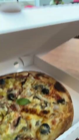 A partially out-of-focus view of a pizza in a white box on a table. Behind the pizza, more tables and chairs are visible, some with red blankets draped over them. The scene appears to be an outdoor seating area of a restaurant. There are trees with hanging lanterns overhead. Buildings are visible in the background.
