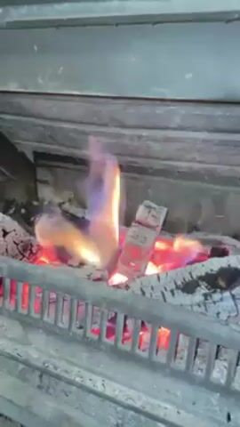 The video shows a fireplace with a metal grate. Behind the grate, there are logs burning with orange, red, and a bit of blue flames. The inside of the fireplace is dark, with gray brick. There is a metallic, gray-colored section above the fireplace opening.