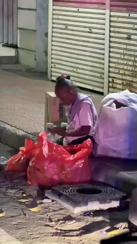 A person is sitting on the sidewalk near a closed store with metal roll-up doors. The person is wearing a light-colored t-shirt and dark pants and has their hair pulled back. They are surrounded by several large plastic bags, including two bright red ones in the foreground and a white one to their right. A cardboard box with the word "ades" visible is behind the person. The person appears to be sorting through items in their hands. The ground is dirty, with debris scattered around. The scene is dimly lit, suggesting it is nighttime or dusk.