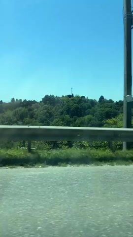 The video captures a roadside view from a moving vehicle on a bright, clear day. In the foreground, the asphalt road surface is visible, followed by a silver metal guardrail. Behind the guardrail, there's a strip of green grass and then dense green trees and foliage. In the mid-ground, a hill covered with abundant green trees extends across the horizon. On top of this hill, a tall, slender communication tower or cell tower is visible. The sky above is a vibrant, clear blue with no clouds. Overhead power lines stretch across the upper portion of the frame. Sunlight is evident, suggesting a sunny day. A road sign briefly appears on the right side of the frame but moves too quickly to be deciphered. No people are visible in the video.
