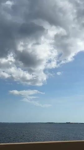 A slightly shaky, vertical, wide shot captures a body of water under a partly cloudy sky. The water is a dark blue and appears calm, with a faint horizon line in the distance. Along the horizon, a faint strip of land is visible, dotted with what appear to be buildings and trees. The sky above is a bright blue, with fluffy white clouds scattered across it. The most prominent cloud formation is a large, dark gray cloud in the upper left quadrant of the frame, contrasting with the lighter white clouds and blue sky. The bottom of the frame shows a tan, textured surface, possibly a railing or barrier. The overall scene suggests a view from a bridge or elevated vantage point overlooking a bay or coastal area. There are no people or discernible actions visible in the video.