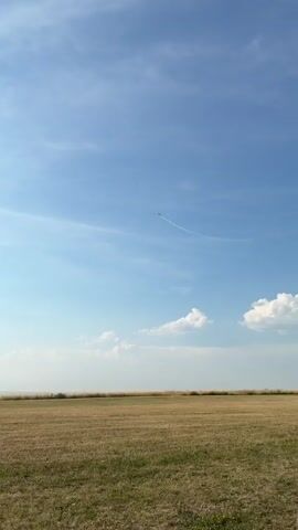 The video shows a small propeller plane performing aerobatics against a clear blue sky. The plane is red and white and leaves a white contrail as it maneuvers. The camera is positioned on the ground, looking upwards. The landscape below consists of a dry, grassy field. The video is short, lasting only about four seconds.