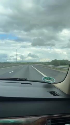 The video is filmed from the interior of a moving car, looking out through the windshield and side mirror. The car is on a highway with multiple lanes and a white line marking the edge of the road. The sky is cloudy with patches of blue visible. There are streetlights on the side of the road and a guardrail. In the side mirror, another car is visible. The landscape outside the car consists of green fields and trees. There is a circular sticker on the windshield.